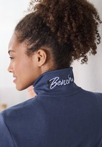 Navyblauwe hoge hals sweatshirt met geborduurd "Bench" logo op de kraag. Zachte stof textuur en een getailleerd ontwerp zichtbaar vanuit het zijaanzicht.