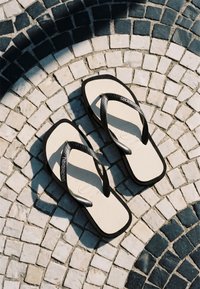 Une paire de tongs Havaianas blanche et noire posée sur un pavement en pierre noir et blanc à motif circulaire.