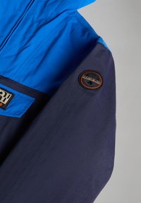 Kleurrijke jas met een blauwe bovenkant en een marineblauwe onderkant. Heeft een logo-opdruk op de mouw en een voorzak met een contrasterend label.
