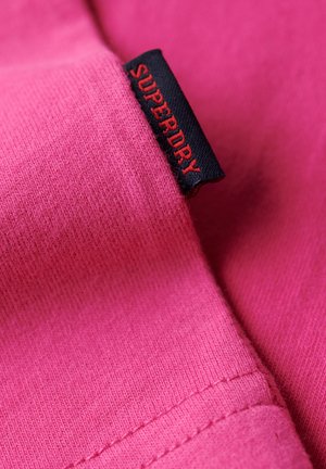 Roze stof close-up met een gestructureerd oppervlak, voorzien van een zwart label met "SUPERDRY" in rode letters. Glad stiksel zichtbaar.