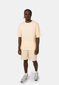 Maglietta a maniche corte a coste color crema e pantaloni corti coordinati, abbinati a scarpe da ginnastica grigie, indossata da un modello che si trova davanti a uno sfondo neutro.