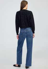 Una donna di spalle indossa un maglione nero a pois, jeans blu a vita alta e ballerine nere su uno sfondo neutro.