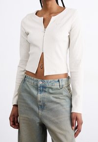 Top corto de manga larga de canalé blanco con cremallera y cuello redondo, combinado con unos vaqueros de denim azul claro. Detalles en hardware plateado en la cremallera.