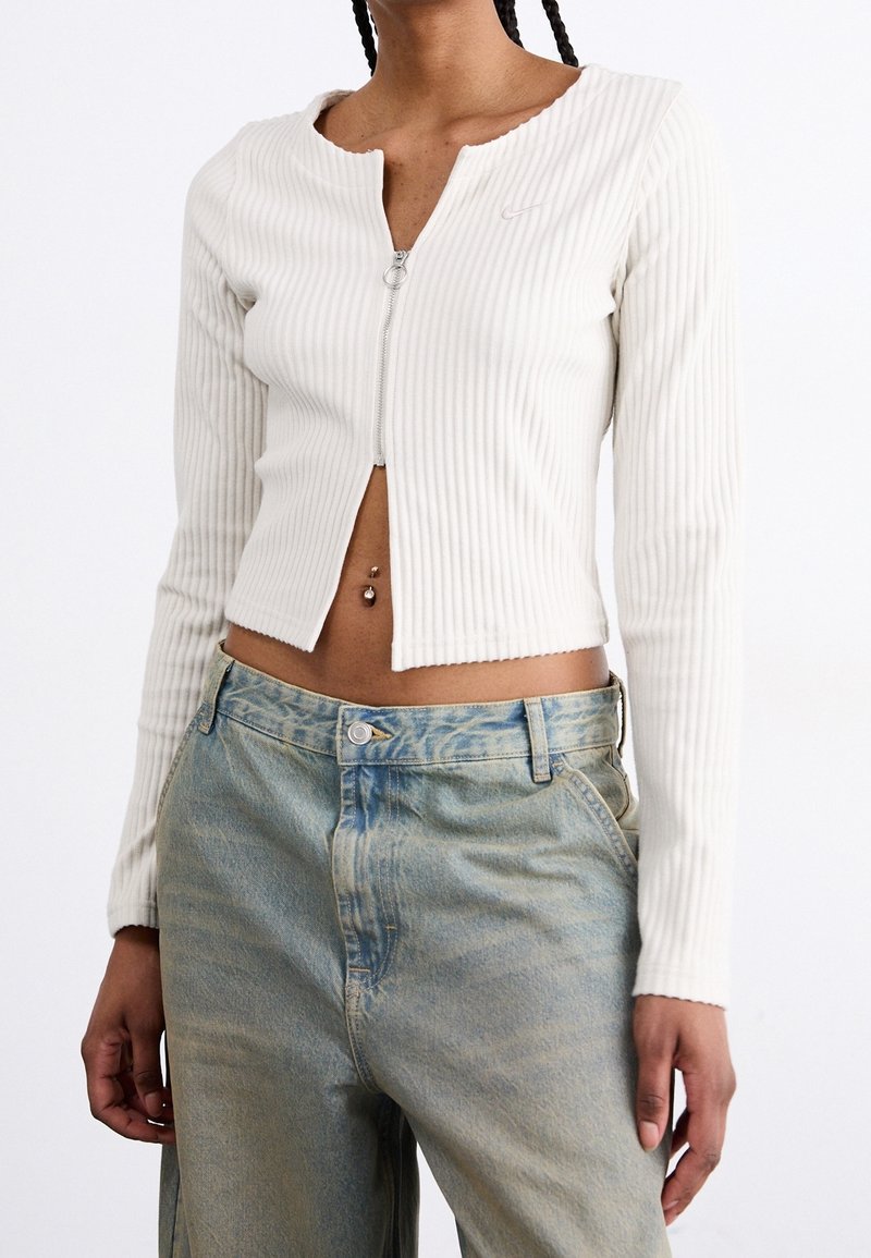 Top corto de manga larga de canalé blanco con cremallera y cuello redondo, combinado con unos vaqueros de denim azul claro. Detalles en hardware plateado en la cremallera.