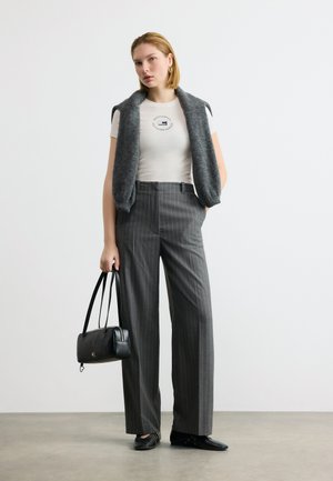 Γυναίκα που φοράει λευκό μπλουζάκι με λογότυπο, γκρι παντελόνι φαρδύ με ρίγες, γκρι αμάνικο πουλόβερ ριγμένο στους ώμους, μαύρα παπούτσια και κρατά μαύρη τσάντα χειρός.