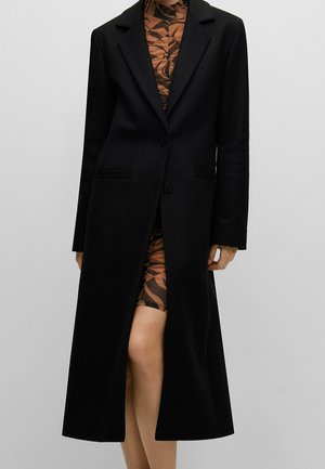 Długi czarny płaszcz o dopasowanym kroju, z kołnierzem w stylu marynarki, dwiema guzikami i rozcięciem z przodu. Wykonany z gładkiego materiału o minimalnej teksturze.