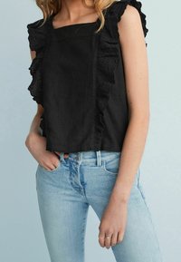 Kvinde iført en sort ærmeløs top med volangdetaljer i blonde og lyseblå højtaljede jeans, hånd hvilende i lommen på jeans.