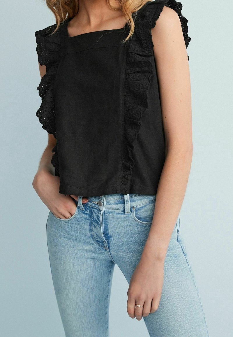 Kvinde iført en sort ærmeløs top med volangdetaljer i blonde og lyseblå højtaljede jeans, hånd hvilende i lommen på jeans.