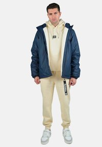 Veste à capuche marine avec doublure en fausse fourrure blanche, sweat à capuche beige et pantalon de jogging assorti. Baskets blanches avec accents gris. Étiquette de marque visible.