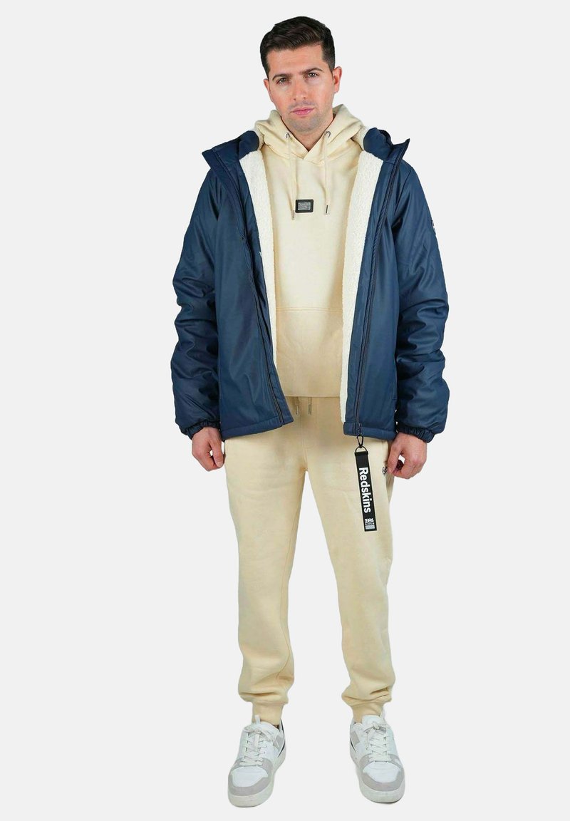 Veste à capuche marine avec doublure en fausse fourrure blanche, sweat à capuche beige et pantalon de jogging assorti. Baskets blanches avec accents gris. Étiquette de marque visible.