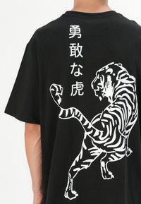 Schwarzes Baumwoll-T-Shirt mit einem großen weißen Grafikdruck eines Tigers und japanischem Text auf dem Rücken, mit lockerer Passform und kurzen Ärmeln.