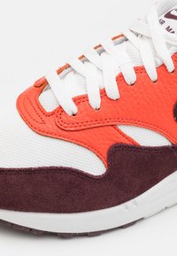 Gros plan d'une basket avec un mesh blanc, des empiècements en cuir orange, des accents en daim marron foncé et des lacets blancs sur une semelle blanche.