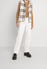 Karierter Wollwesten in Weiß, Grau und Braun, kombiniert mit einem braunen gerippte Pullover und weißen geraden Hosen. Schwarze Plateauschuhe runden das Outfit ab.