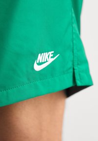 Zelené šortky Nike s hladkým povrchem, s bílým vyšitým logem poblíž lemu. Design zahrnuje jednoduchý střih a čisté okraje.