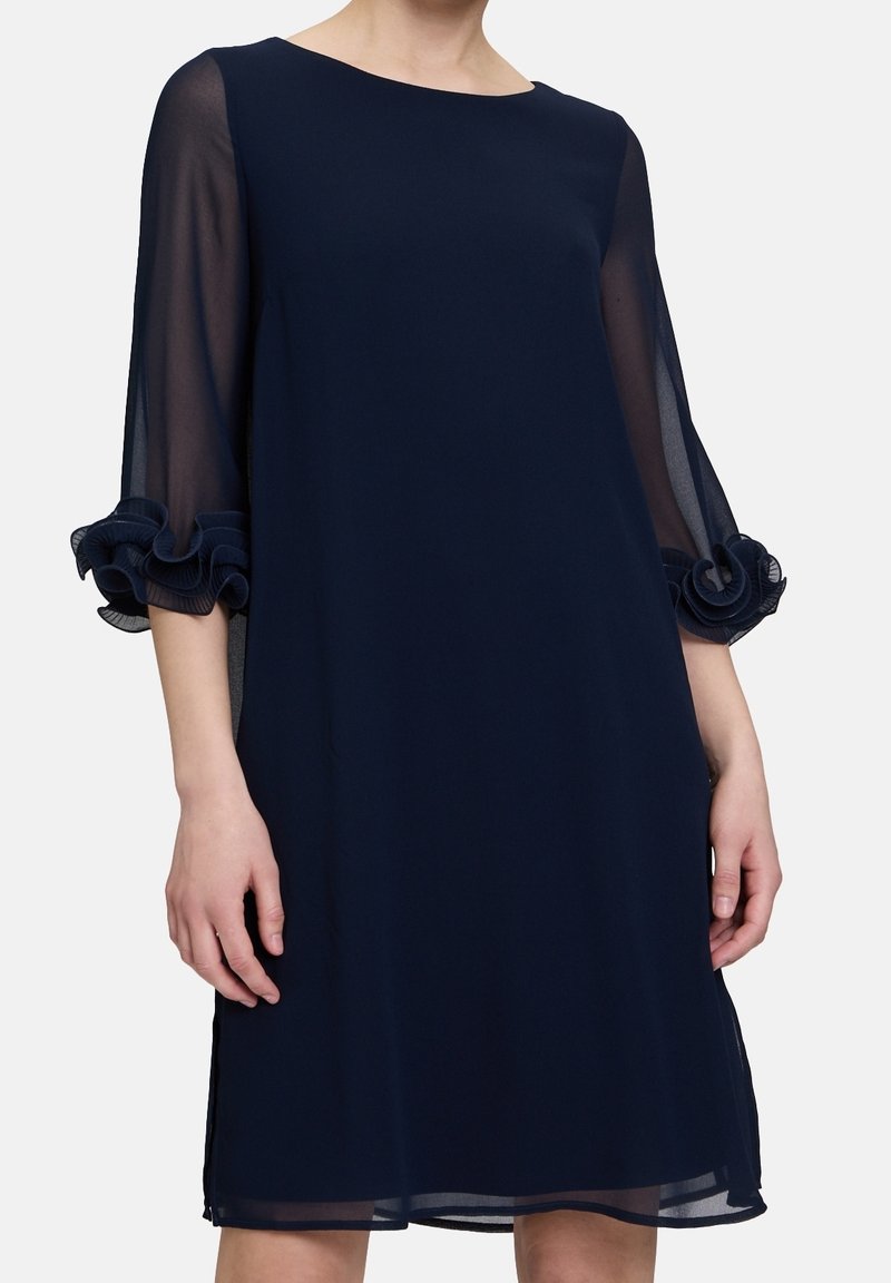 Femme portant une robe bleu marine longueur genou avec des manches trois-quarts transparentes et des poignets à volants, debout devant un fond clair uni.