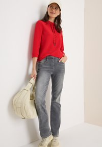 Rode geribbelde trui met trekkoordjes, grijze jeans en beige tas. Model draagt lichtgekleurde sneakers. Eenvoudige, casual outfit tegen een neutrale achtergrond.