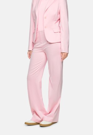 Woman wearing a light pink suit with a fitted blazer and wide-leg trousers paired with white sneakers on a plain background.