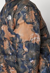 Ocieplana kurtka z wielokolorowym, brązowym i niebieskim abstrakcyjnym wzorem. Posiada logo na górnej lewej stronie, z widocznymi szwami i fakturą.