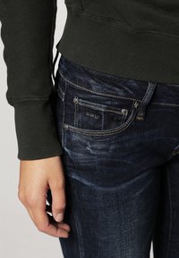 Mörkblå denimjeans med subtila blekningar, fickdetaljer och guldstikningar, tillsammans med en mörkgrön ribbad sweatshirtärm.