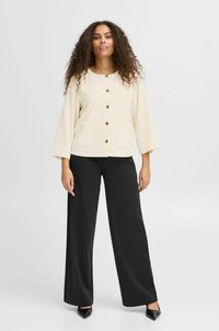 Chaqueta beige de botones con tejido texturizado, dos bolsillos frontales y mangas anchas, combinada con pantalones de pierna ancha negros y zapatos de punta negros.