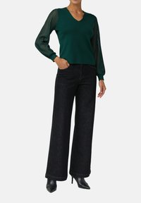 Frau trägt einen dunkelgrünen V-Ausschnitt-Pullover mit transparenten gestreiften Ärmeln, schwarze weit geschnittene Jeans und schwarze spitze Stiefeletten mit Absatz.