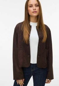 Mlada žena s dugom ravnom kosom koja nosi tamnosmeđu kaput s dugmadi, bijelu košulju i tamnoplave traperice nasuprot jednolične pozadine.