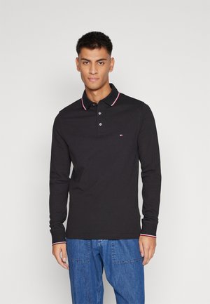Camisa polo negra de manga larga con acentos en el cuello en rojo y blanco, que presenta una tapeta de tres botones y un parche de logo en el lado izquierdo del pecho.