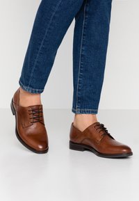 Chaussures habillées en cuir marron avec une finition lisse, dotées d'un bout pointu, de lacets et d'un petit talon, associées à un jean en denim bleu foncé.