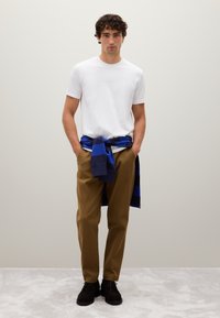 Uomo in piedi con le mani in tasca, che indossa una maglietta bianca, pantaloni marroni, scarpe nere e un maglione a righe blu-neri annodato in vita.