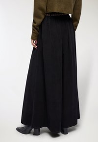 Gonna maxi nera plissettata con una texture liscia, dotata di una vita accentuata da una cintura con borchie. Indossata con stivaletti con tacco.
