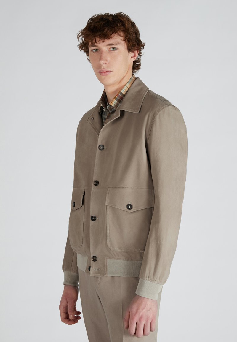 Giacca marrone chiaro realizzata in tessuto liscio, con chiusura a bottoni, due tasche frontali e polsini a coste. Il modello è in piedi con un'espressione neutra.