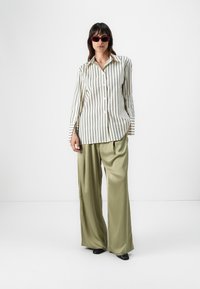 Striped button-up shirt in cream and black, paired with olive green wide-leg pants made of smooth fabric. Accessorized with dark sunglasses.