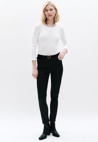 Witte geribbelde trui, zwarte aansluitende broek en enkellaarsjes. De outfit bevat een kettinghalsband en een ronde riemsluiting.
