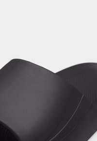 Κοντινή λήψη μιας μαύρης παντόφλας με ανάγλυφη επιφάνεια στο πέλμα και φαρδιά λωρίδα στο πάνω μέρος, σε απλό φόντο.