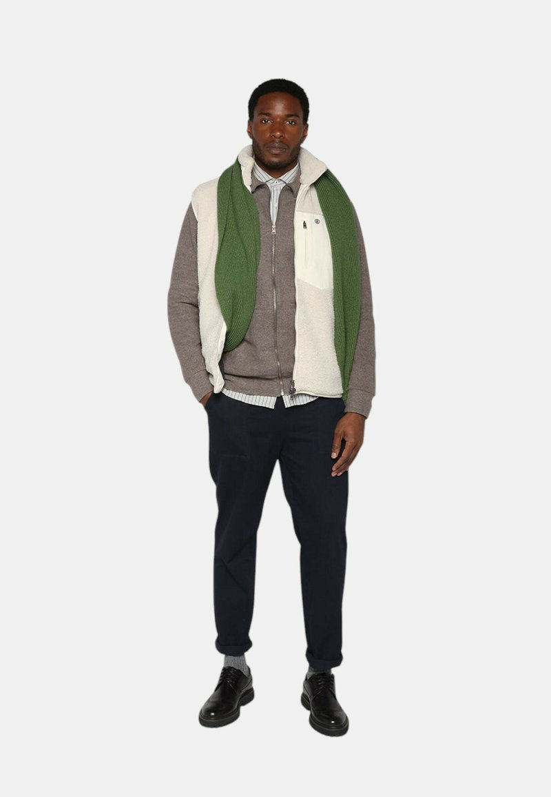 Tenue d'homme comprenant un gilet en matériaux mélangés blanc et vert sur un pull gris, un pantalon bleu marine et des chaussures noires avec des chaussettes grises.