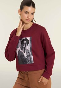 Felpa borgogna con design accorciato, dotata di una stampa grafica in bianco e nero di una donna con il testo 'IL MOVIMENTO È UNA TERAPIA.' Tessuto morbido.