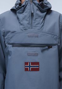 Lichtblauwe, waterdichte anorak met een met rits sluitende borstzak, klittenbandaccenten en een geborduurd Noors vlagpatch in rood, blauw en wit.
