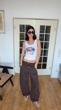 Νεαρή γυναίκα με γυαλιά ηλίου, λευκό αμάνικο μπλουζάκι με σχέδιο, φαρδιά καφέ παντελόνια και μεταλλικά αθλητικά παπούτσια, στέκεται μέσα κοντά σε διπλές πόρτες με γυάλινα πάνελ.