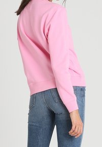 Felpa rosa con scollo a girocollo, polsini e orlo a costine. Il materiale sembra morbido e liscio, indossata con jeans di denim blu.