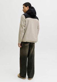 Veste bicolore beige et noire avec capuche, présentant une texture lisse et une coupe décontractée. Portée avec un jean ample foncé et des chaussures beige.