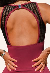 Maillot de bain bordeaux avec un dos en maille noire, design à découpe courbée, fermoir en argent en haut et tissu texturé. Mains reposant sur les hanches.
