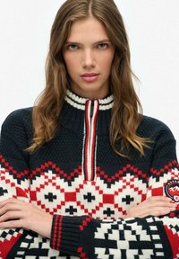 Marineblauwe, rode en witte gebreide trui met een hoge kraag, ritssluiting, geometrische patronen en contrasterende ribbelmanchetten. Heeft een logo-patch op de mouw.