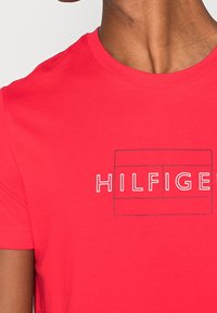 Camiseta de algodón roja con un estampado gráfico que incluye el texto "HILFIGER" dentro de un contorno rectangular. Cuello redondo y mangas cortas.