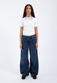 Vit åtsittande t-shirt i kombination med högmidjade, vida mörkblå denimjeans. Modellen bär svarta skor och lager av silverhalsband.