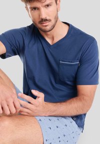 Camiseta de cuello en V azul marino con un bolsillo frontal, combinada con pantalones cortos azul claros con un pequeño patrón de palmeras. Tejido suave, corte relajado.