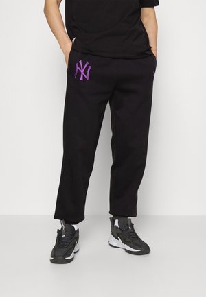 Personne portant un pantalon de survêtement noir avec un logo New York violet sur la cuisse, des baskets noires et des chaussettes blanches, debout les mains dans les poches.