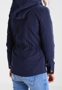 Granatowa kurtka z kapturem z taliowaniem, wykonana z gładkiego materiału, z srebrnymi zatrzaskami na rękawach. Częściowy widok od tyłu.