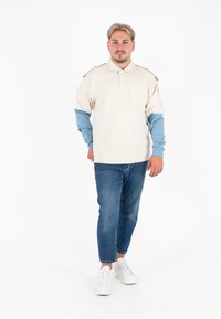 Korte mouwen polo-shirt in crème met blauwe mouwen, zak op de borst en contrasterende stiksels, gecombineerd met slim-fit blauwe jeans en witte sneakers.