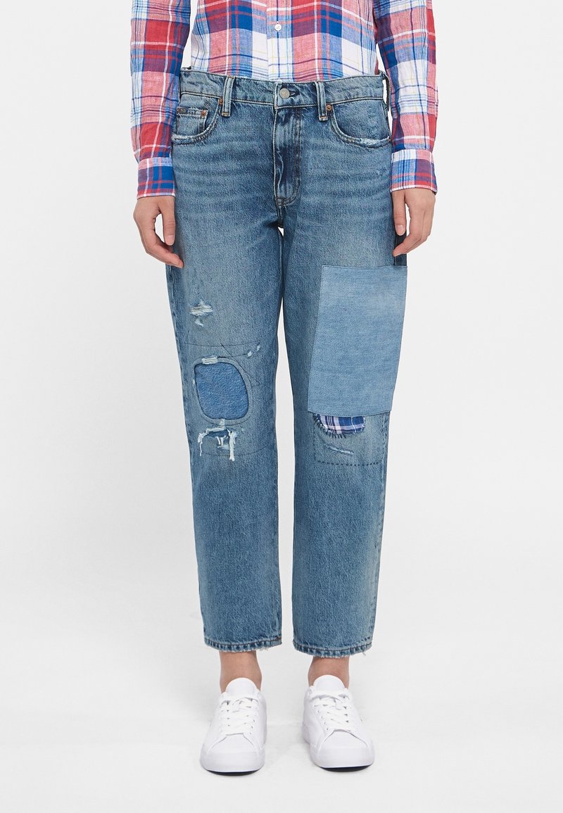 Denim jeans in light blue with a relaxed fit. Features distressed areas and patch details in darker shades of blue. White sneakers complement the look.