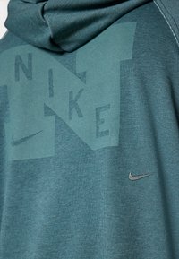 Tamsiai žalia kapučino sportinė palaidinė su dideliu toniniu "NIKE" grafiniu logotipu ir mažuoju sidabriniu Swoosh logotipu apatiniame šone. Minkšta tekstūra.